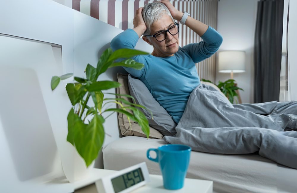 Agitação noturna no idoso. Foto de uma senhora de óculos, sentada na cama com dificuldades para dormir.