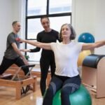 Foto de uma instrutora de pilates ajudando uma senhora no equilibrio na bola.