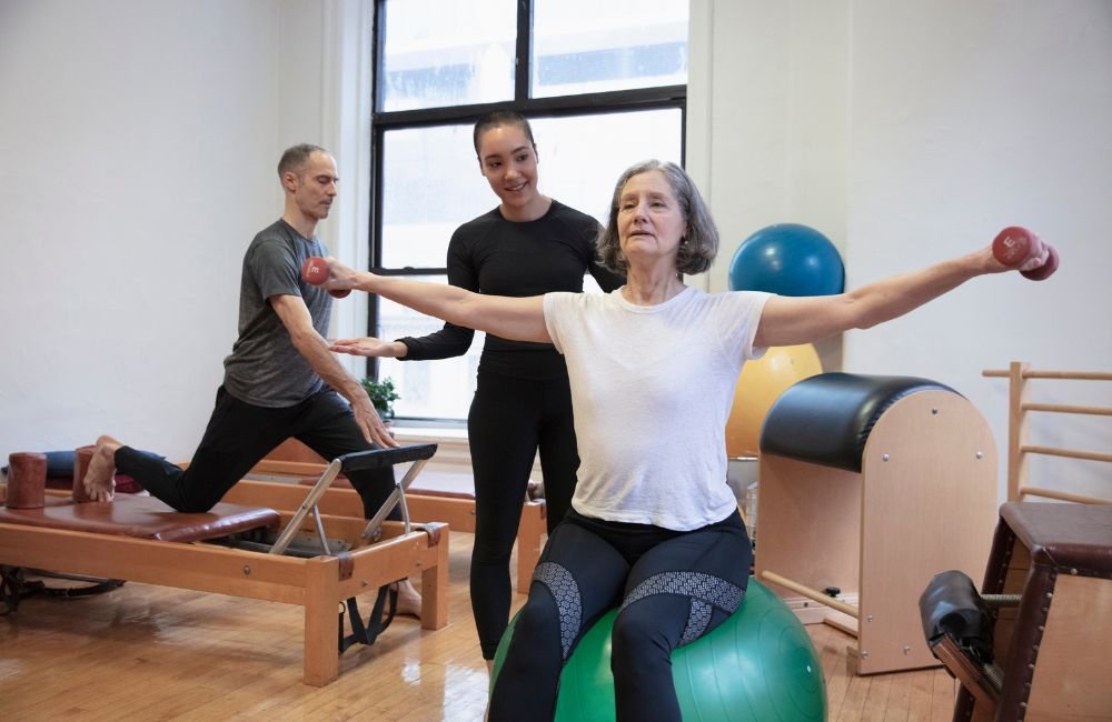 Foto de uma instrutora de pilates ajudando uma senhora no equilibrio na bola.