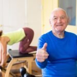 Foto com pesssoas praticando pilates e um senhor idoso fazendo um positivo.