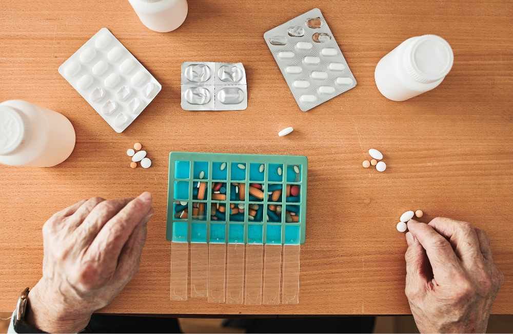 Importante garantir que a medicação seja gerenciada de maneira eficaz. A foto mostra as mãos de uma pessoa arrumando a medicação numa caixinha separadora.